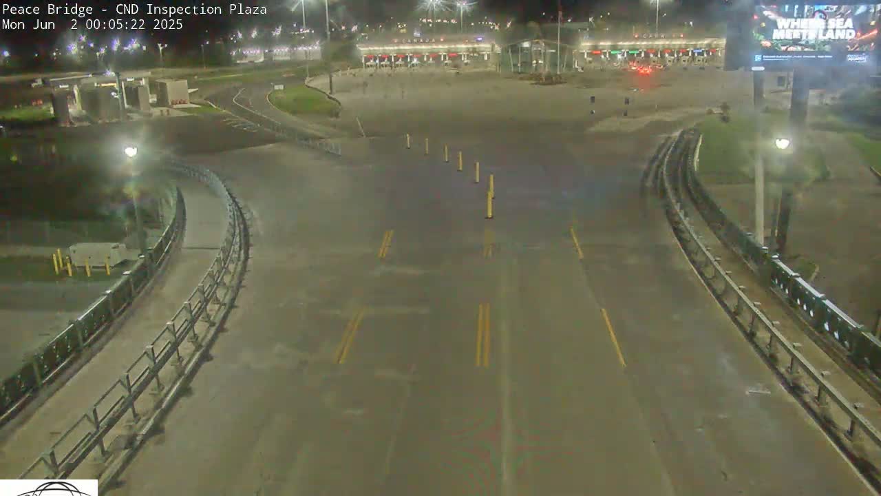 A nighttime, overhead view shows an empty roadway curving toward a border inspection plaza with several yellow traffic markers and minimal lighting.