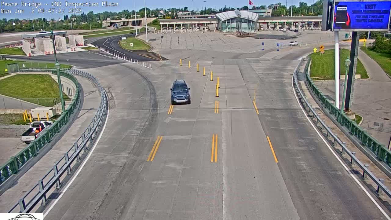A dark-colored SUV drives toward a border inspection plaza on a sunny day.
