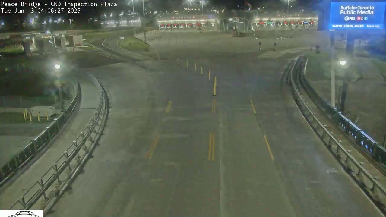 A nighttime, empty roadway curves toward a border inspection plaza under clear skies.