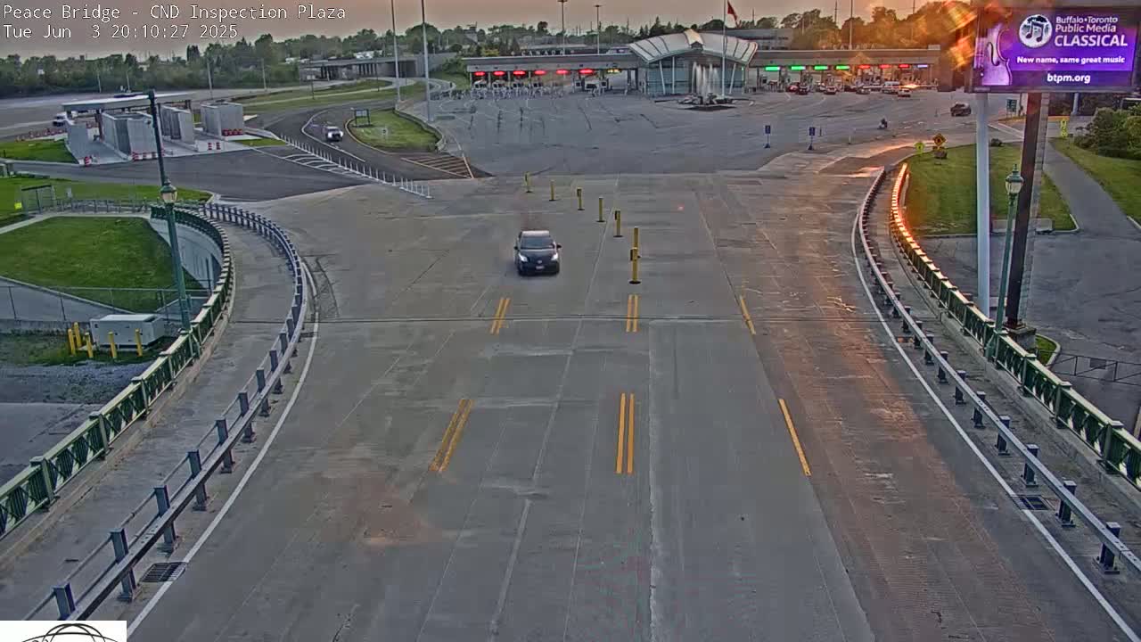 A single dark-colored car drives toward a border inspection plaza at sunset on a paved roadway with green railings.