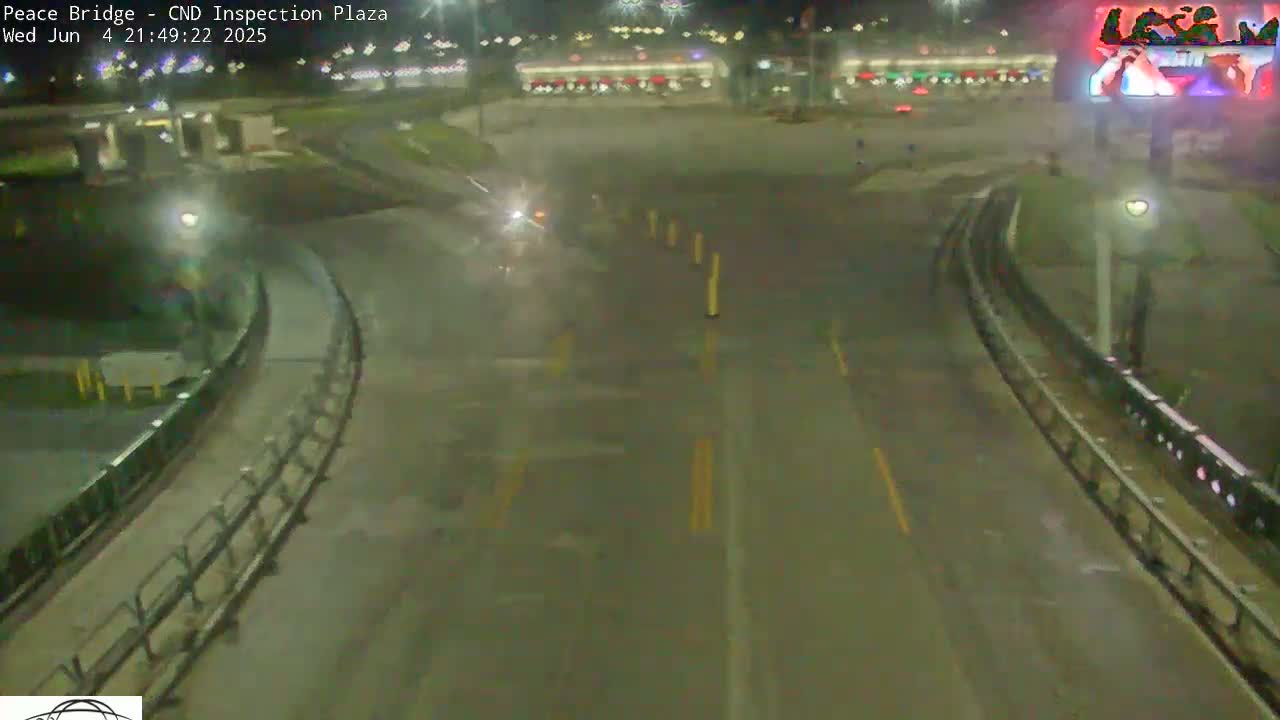 A nighttime, wet roadway curves through a plaza with sparse vehicle traffic under dim lighting.