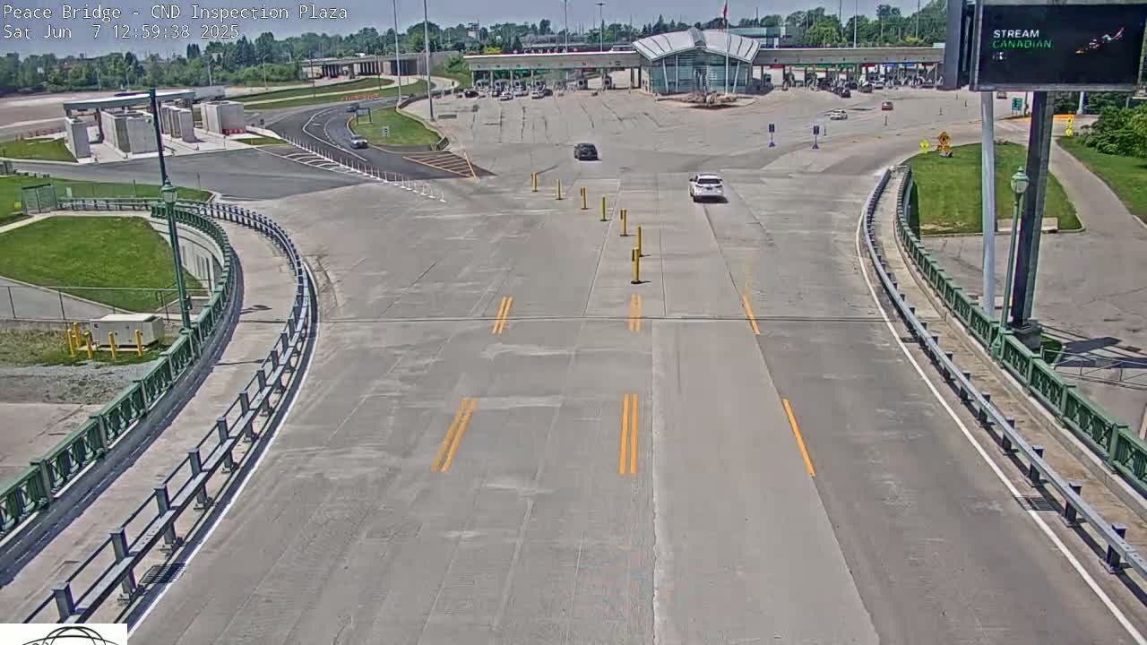 A mostly empty roadway curves toward a border inspection plaza under a sunny sky.