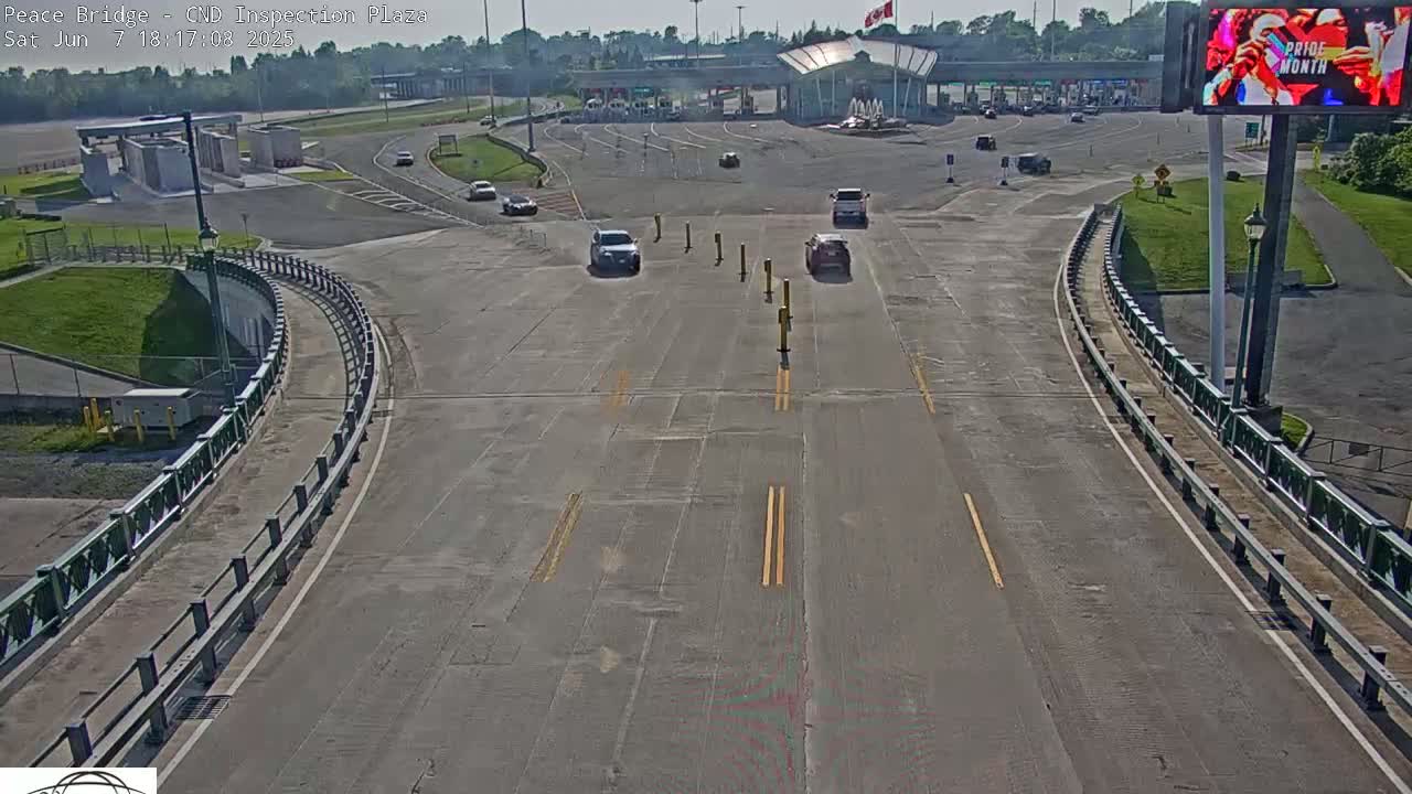 A sunny overhead view of a roadway curving toward a border crossing inspection plaza with several cars visible.