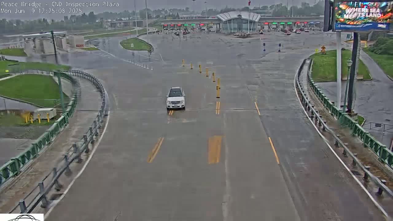 A white SUV drives on a wet roadway toward a border inspection plaza under an overcast sky.