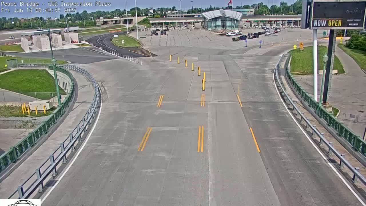 An empty roadway curves toward a border crossing inspection plaza under a sunny sky.
