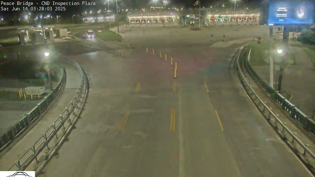 A nighttime, overhead view of a mostly empty roadway leading to a border inspection plaza, illuminated by artificial lights.