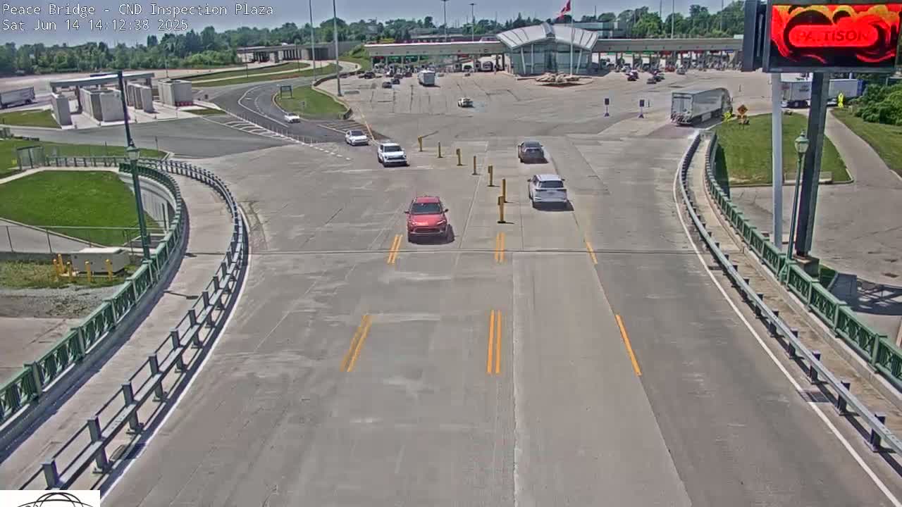 A sunny day at a border crossing inspection plaza shows several cars slowly approaching inspection booths, with a few other vehicles already at the booths or parked nearby.