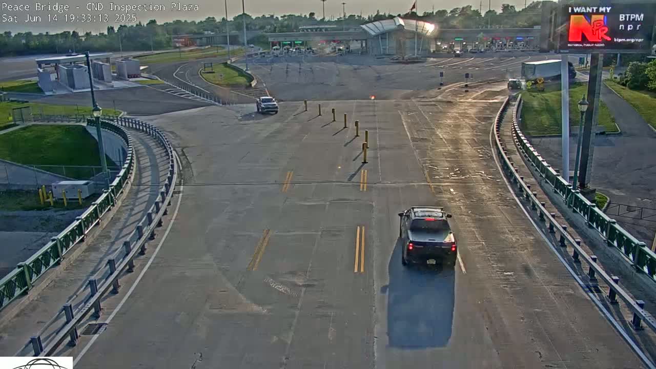A sunny day at a border crossing inspection plaza shows several cars slowly approaching inspection booths, with a few other vehicles already at the booths or parked nearby.