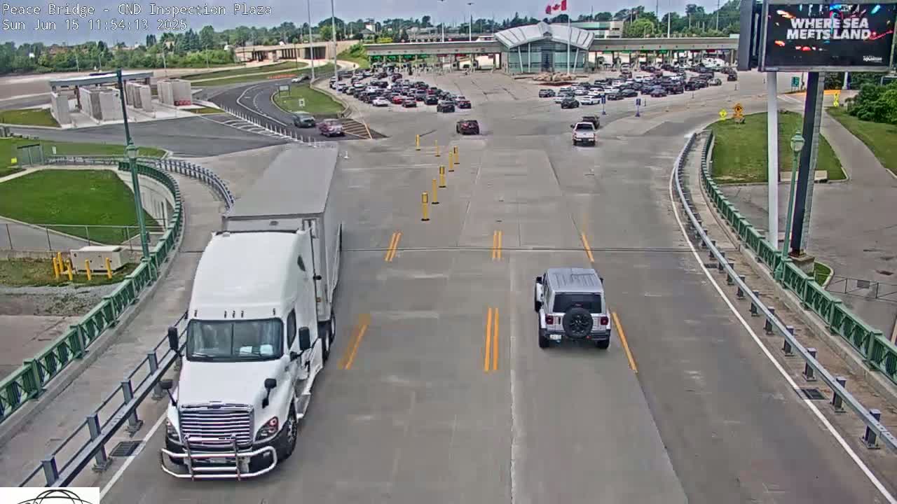 A white semi-truck and a silver SUV are driving on a highway leading to a border crossing plaza under a partly cloudy sky.