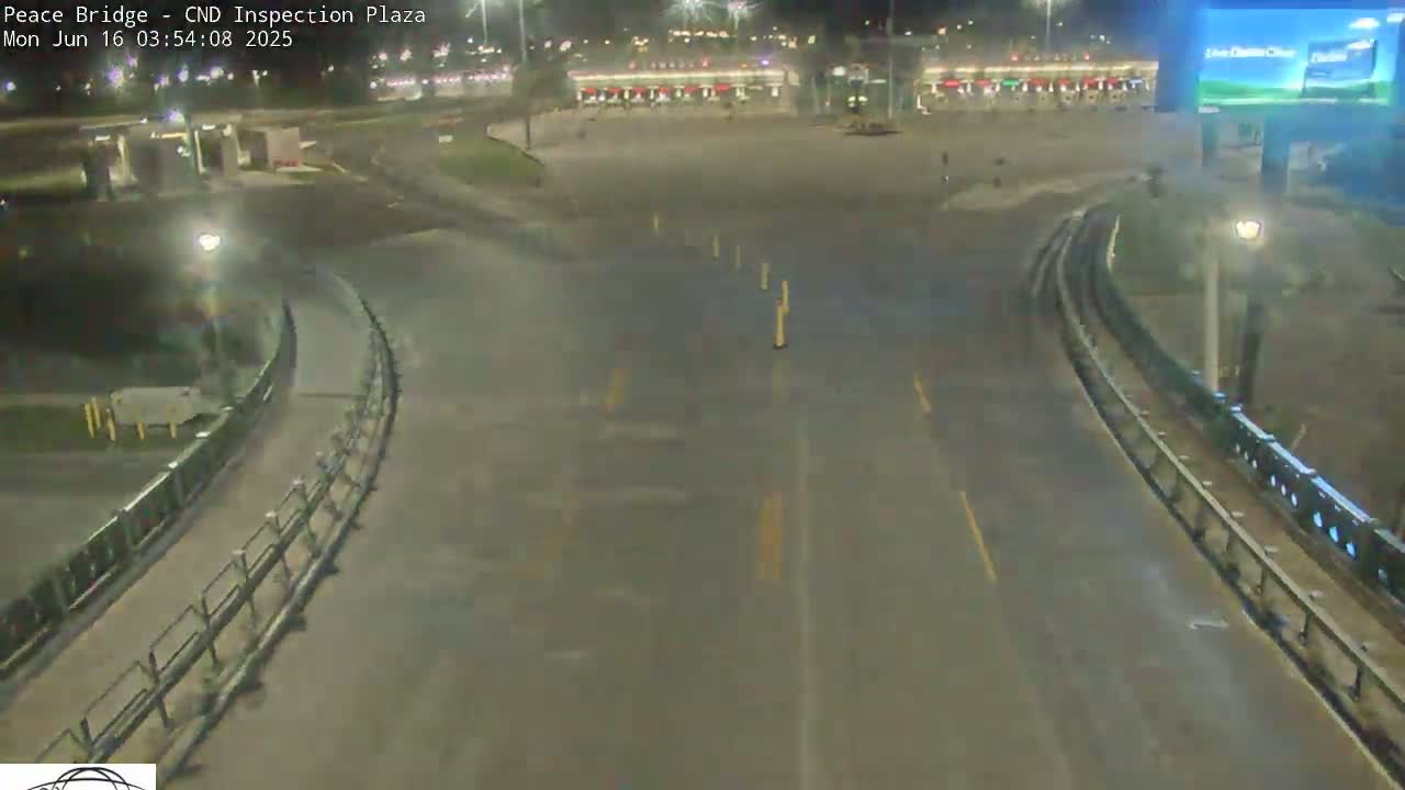 A nighttime, wet roadway curves through a mostly empty inspection plaza with buildings and lights visible in the distance.