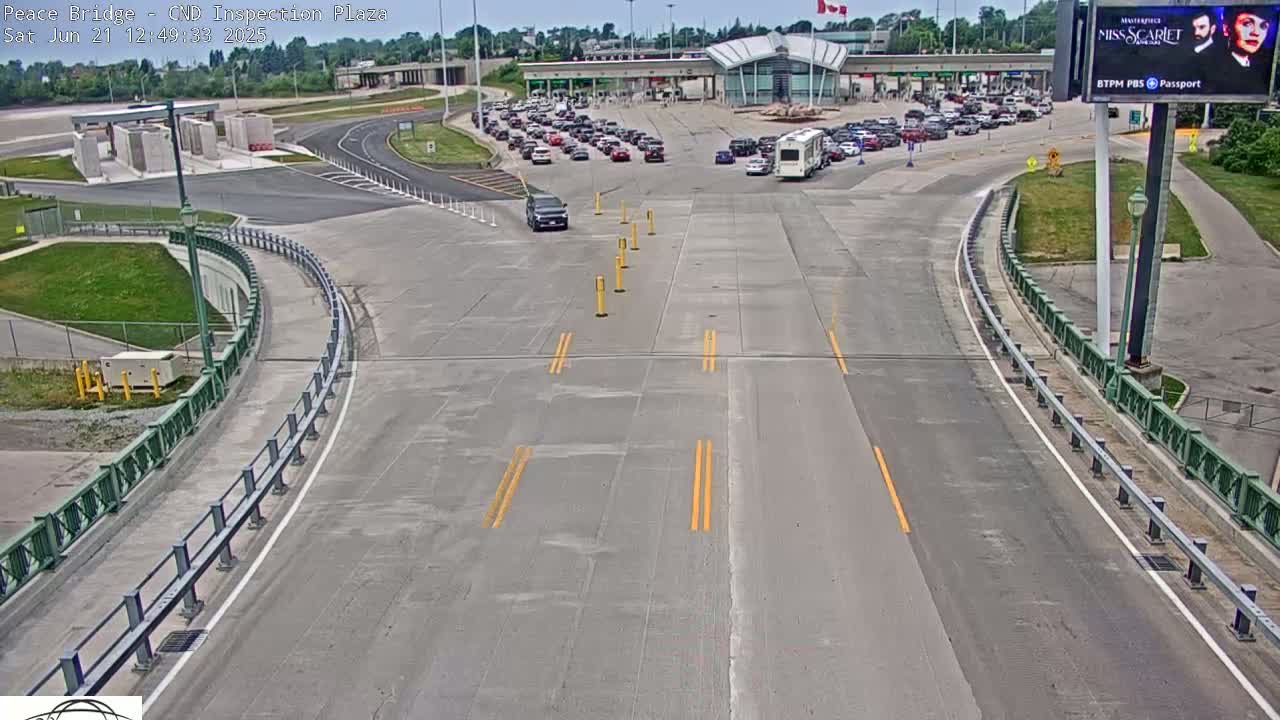 A mostly empty highway approaches a border crossing with several cars waiting in lines on a sunny day.