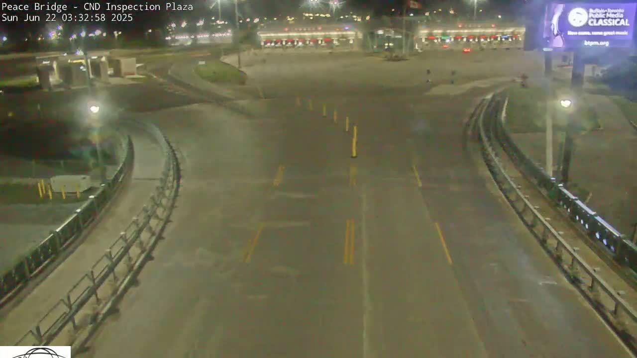 A wet, empty roadway curves through a nighttime border inspection plaza with several buildings visible in the background.