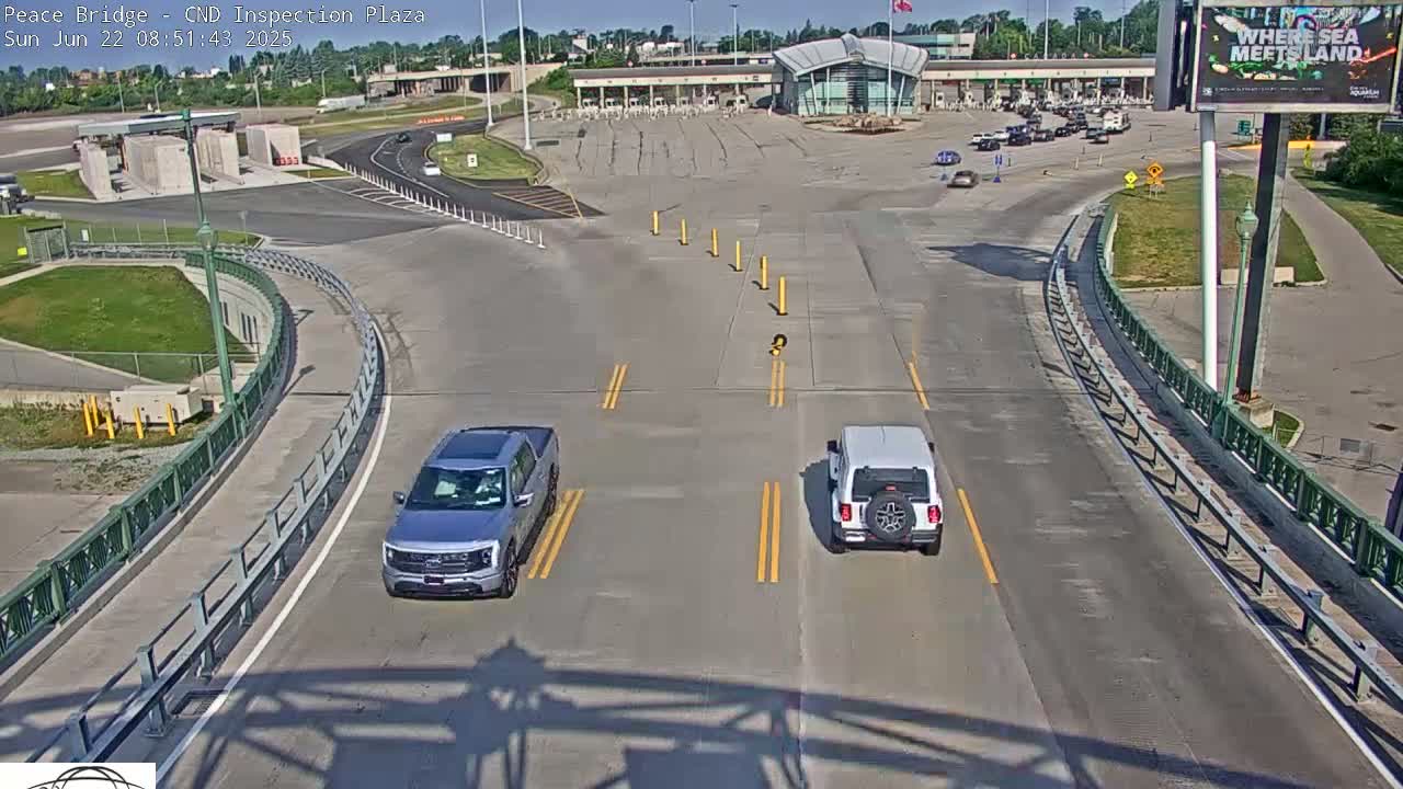 A sunny overhead view of a highway intersection near a border inspection plaza, with two vehicles driving on the roadway.