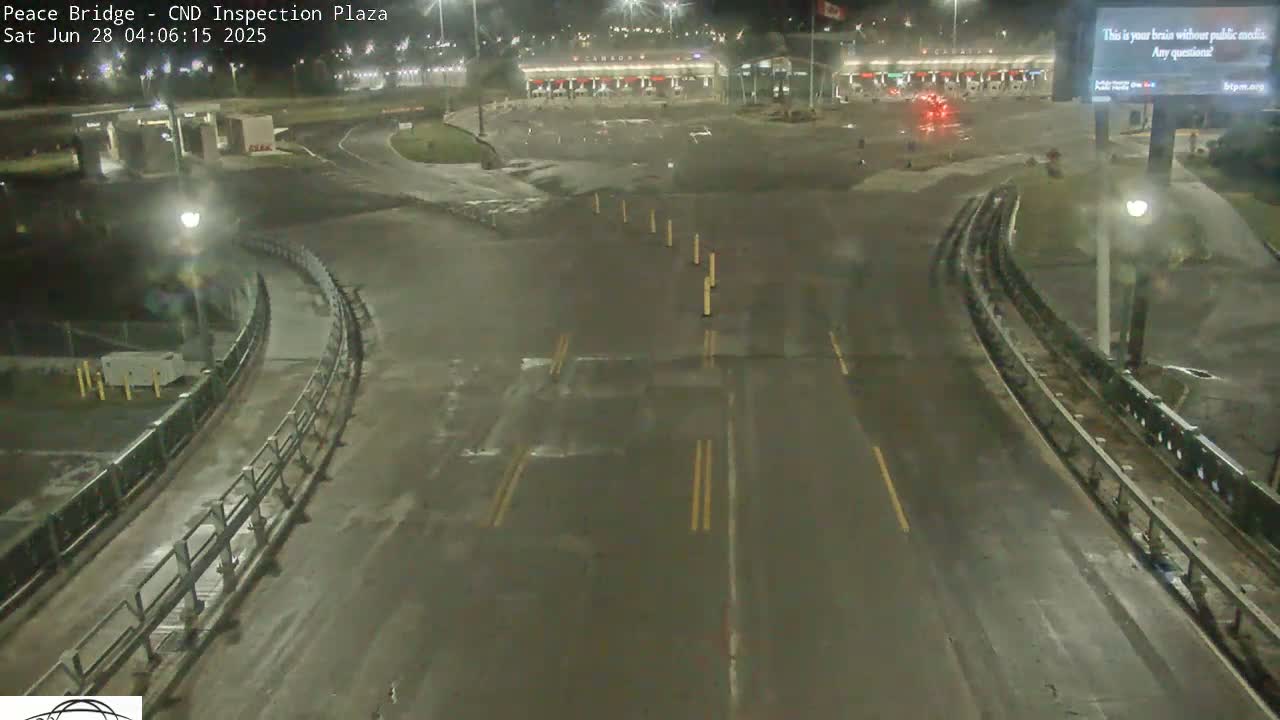 A wet, nighttime view of an empty border crossing inspection plaza with a curving roadway leading to the inspection booths.