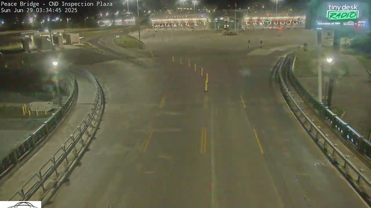A nighttime, empty highway curves toward a border inspection plaza under partly cloudy skies.