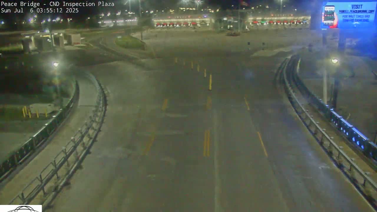 A nighttime view of a mostly empty roadway curving through a plaza with some lighting and barriers, under clear skies.
