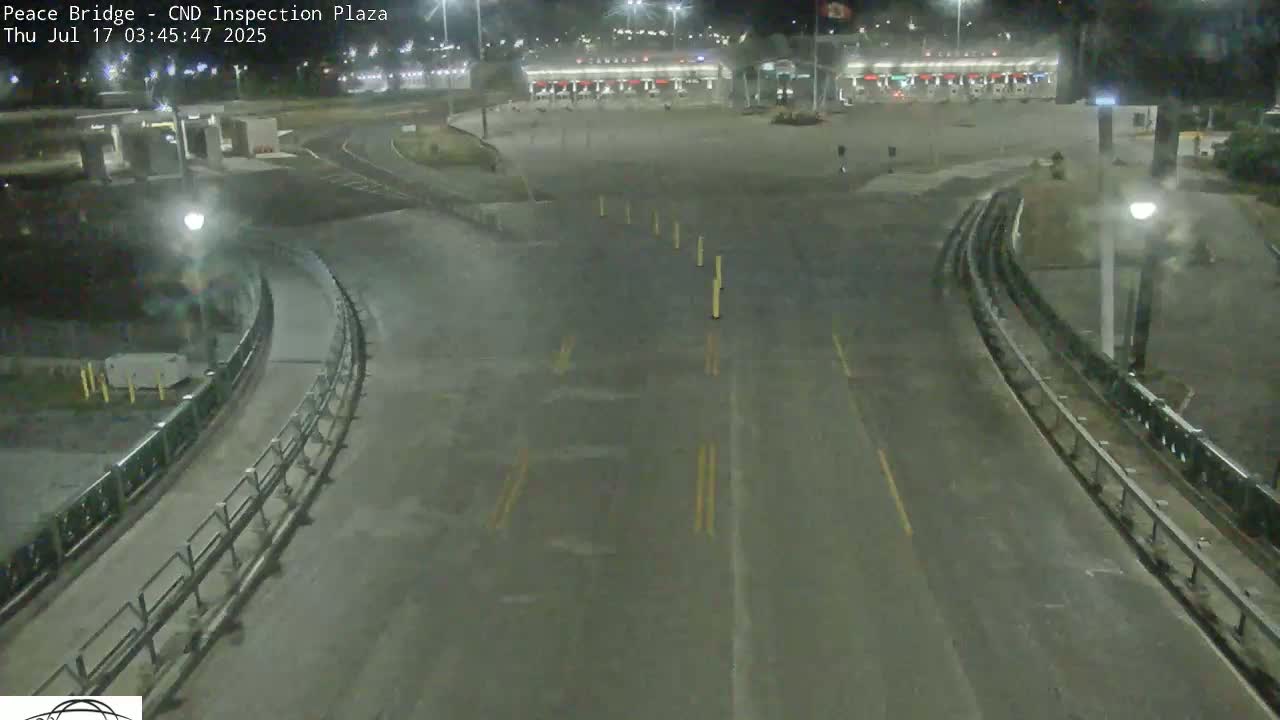 A nighttime, empty roadway curves toward a border inspection plaza under artificial light.