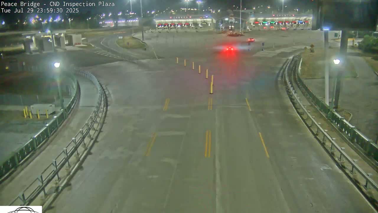 A nighttime, empty roadway curves toward a border inspection plaza under artificial light.