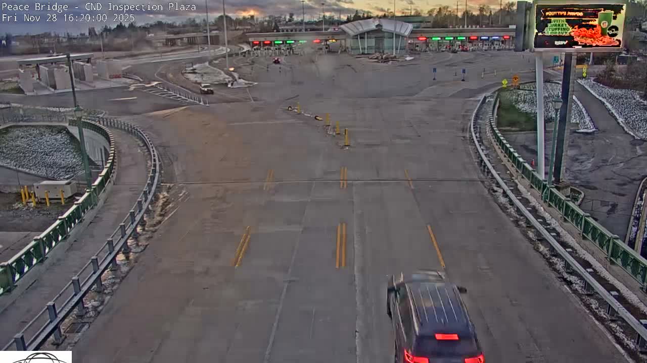A dark SUV travels on a multi-lane road towards a border inspection plaza with multiple entry gates, under a partly cloudy, late afternoon sky with scattered snow on the ground.