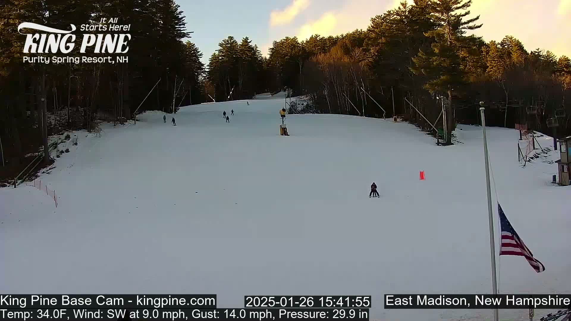 A snow-covered ski slope with several skiers is visible under a partly cloudy sunset sky with a southwest wind.