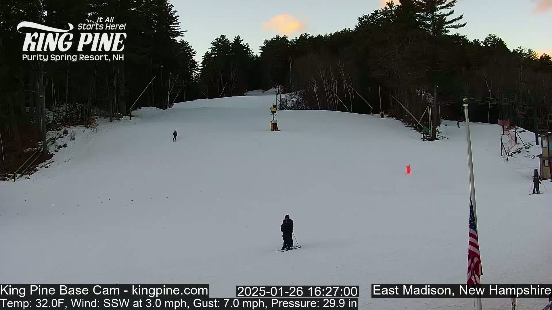 A snowy ski slope at dusk is seen with several skiers, a ski lift, and a small American flag.