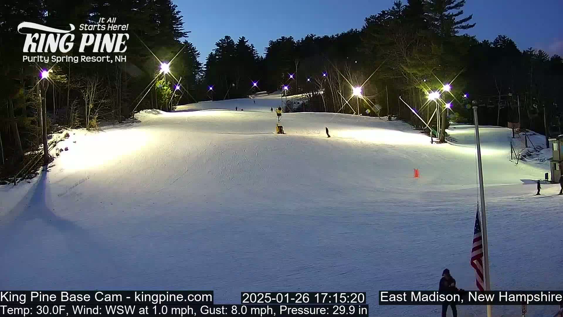 A snow-covered ski slope is illuminated at night by several lights, with a few skiers visible, under a twilight sky.