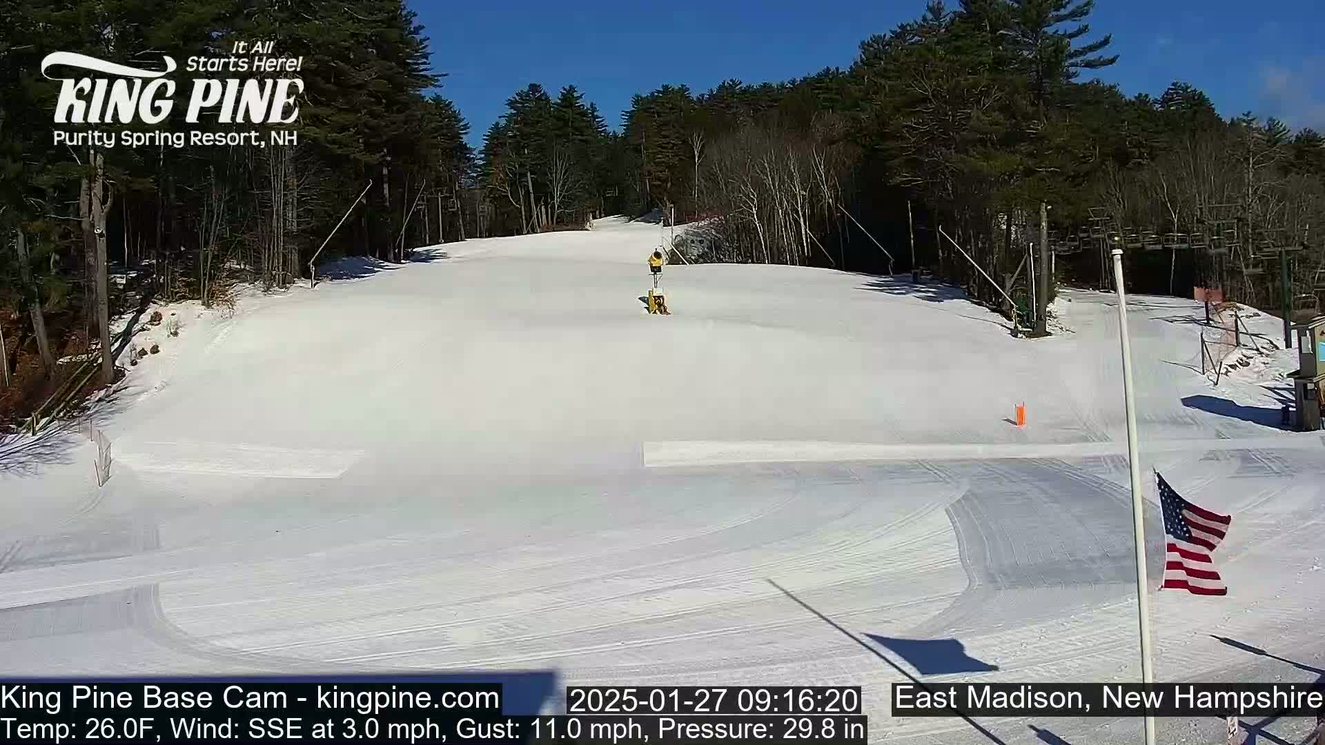 A snow-covered ski slope is visible under a sunny sky, with a person operating a snow groomer in the middle ground and an American flag in the foreground.