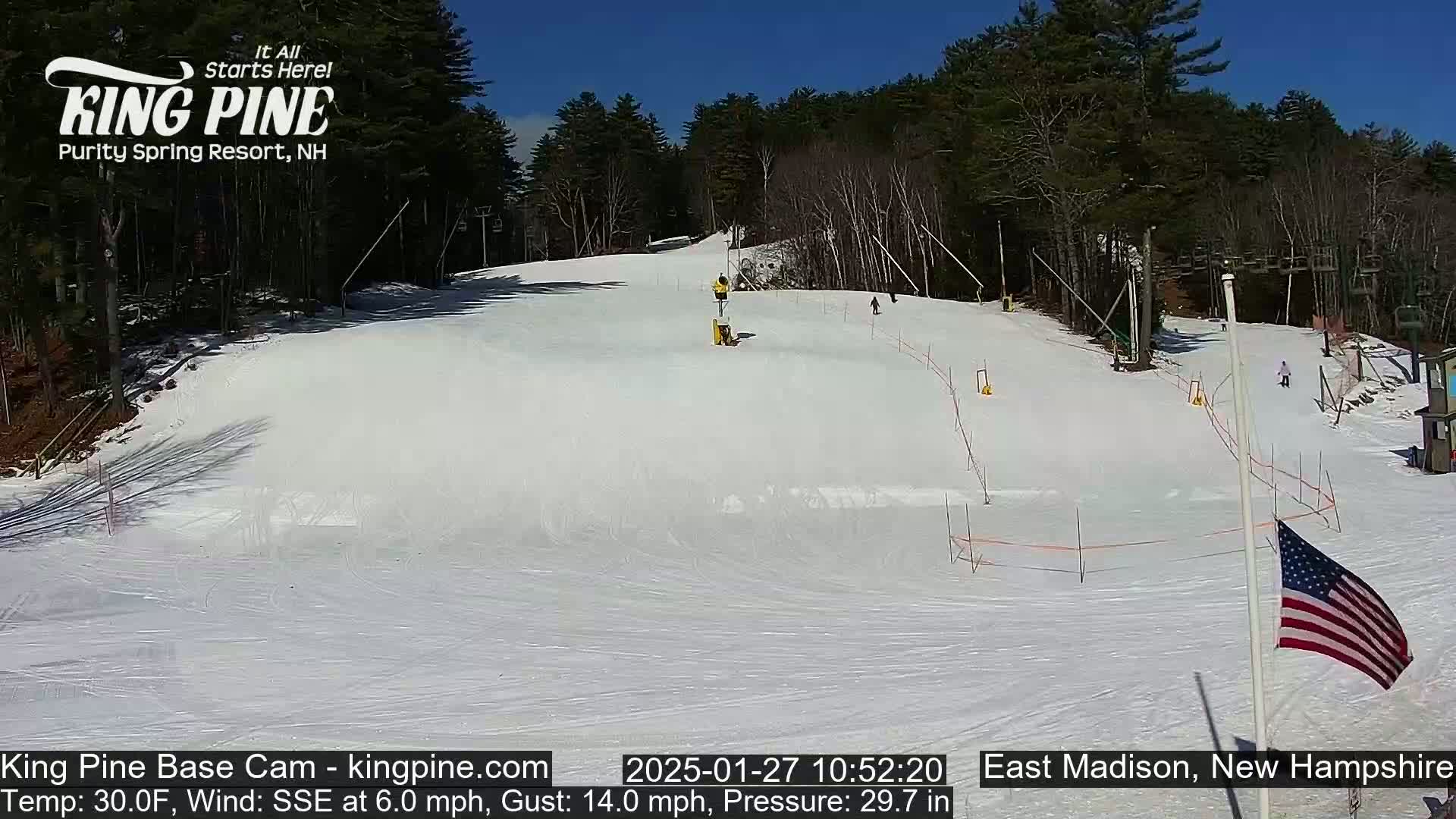 A snow-covered ski slope with several skiers, ski lifts, and an American flag, under a sunny sky.