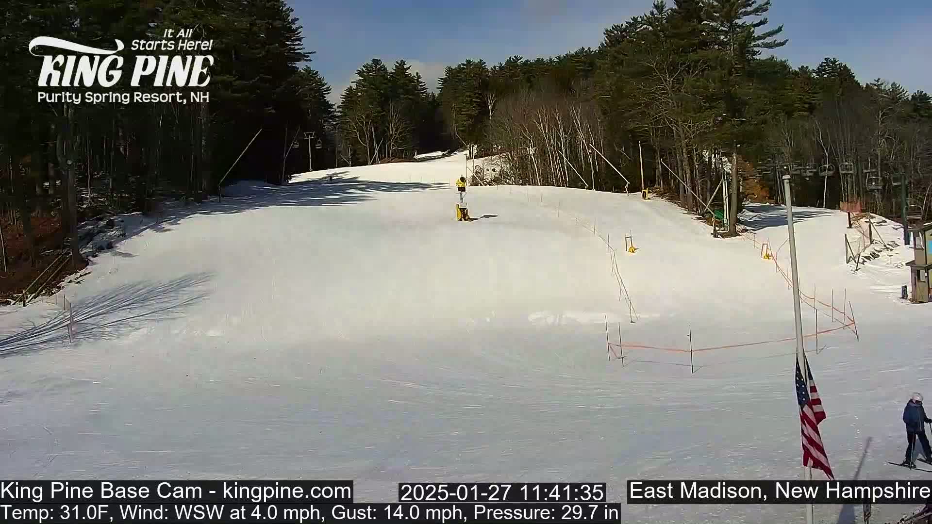 A snow-covered ski slope is visible on a sunny day with a person skiing in the distance, surrounded by trees and ski lifts.