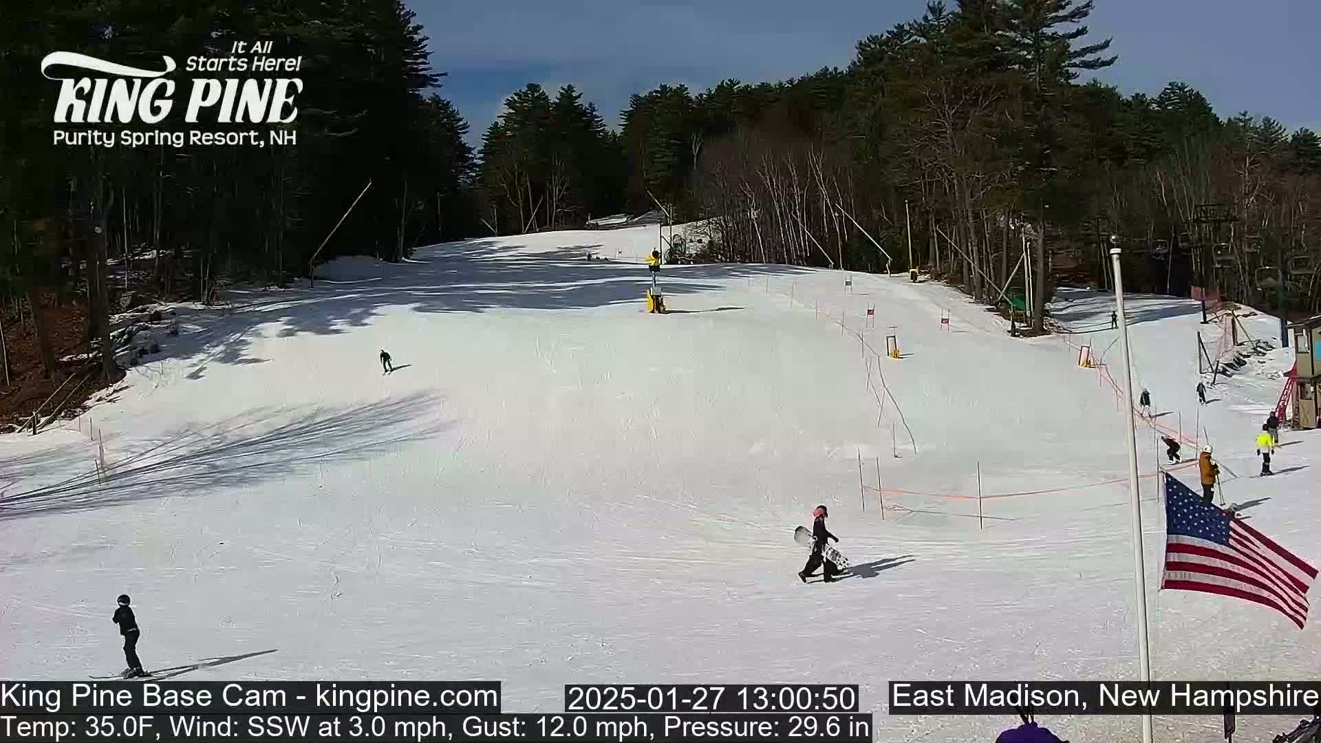 A sunny snow-covered ski slope with several skiers and snowboarders, surrounded by trees.