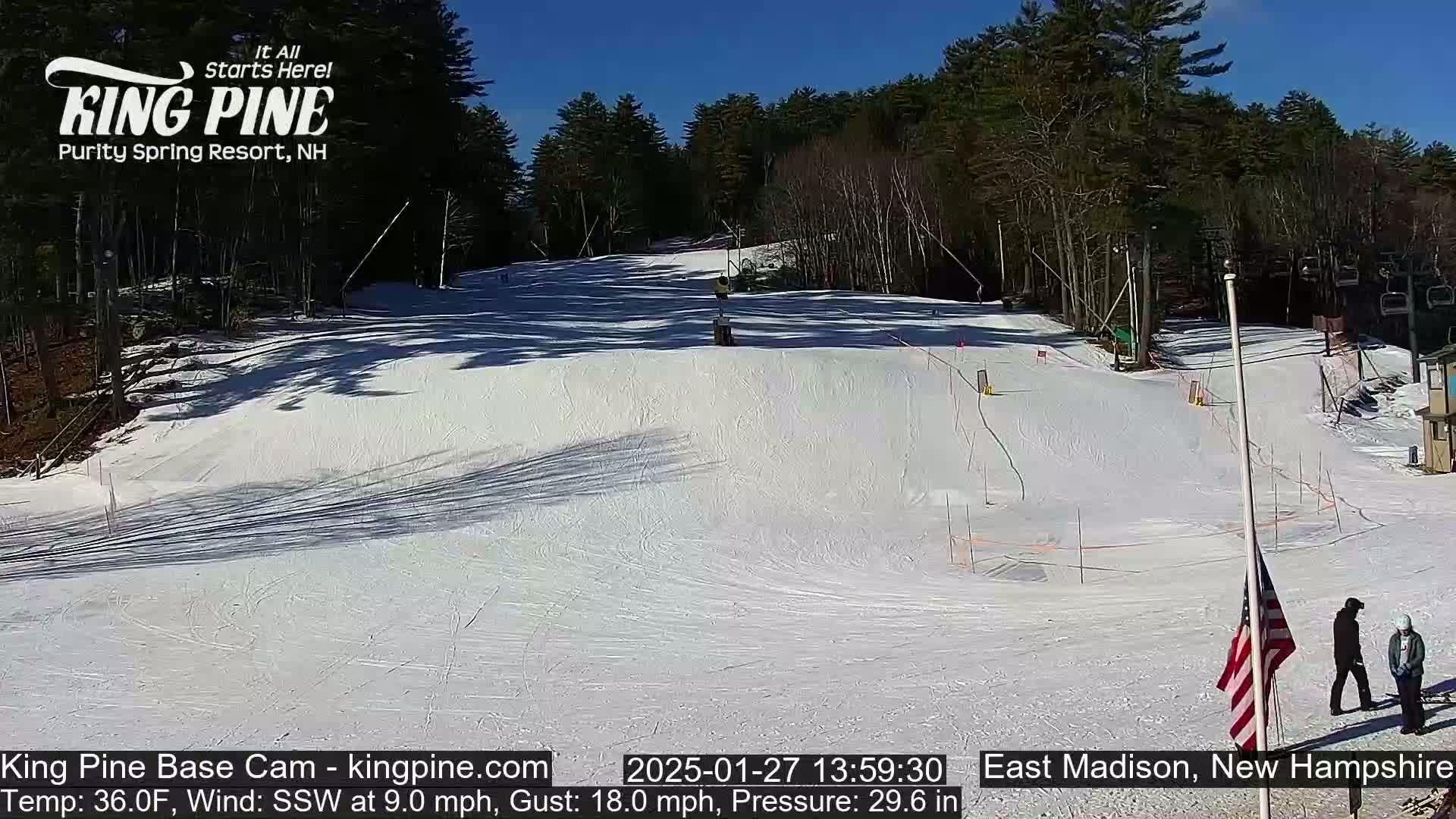 A snow-covered ski slope on a sunny day, with two people standing near an American flag at the bottom.