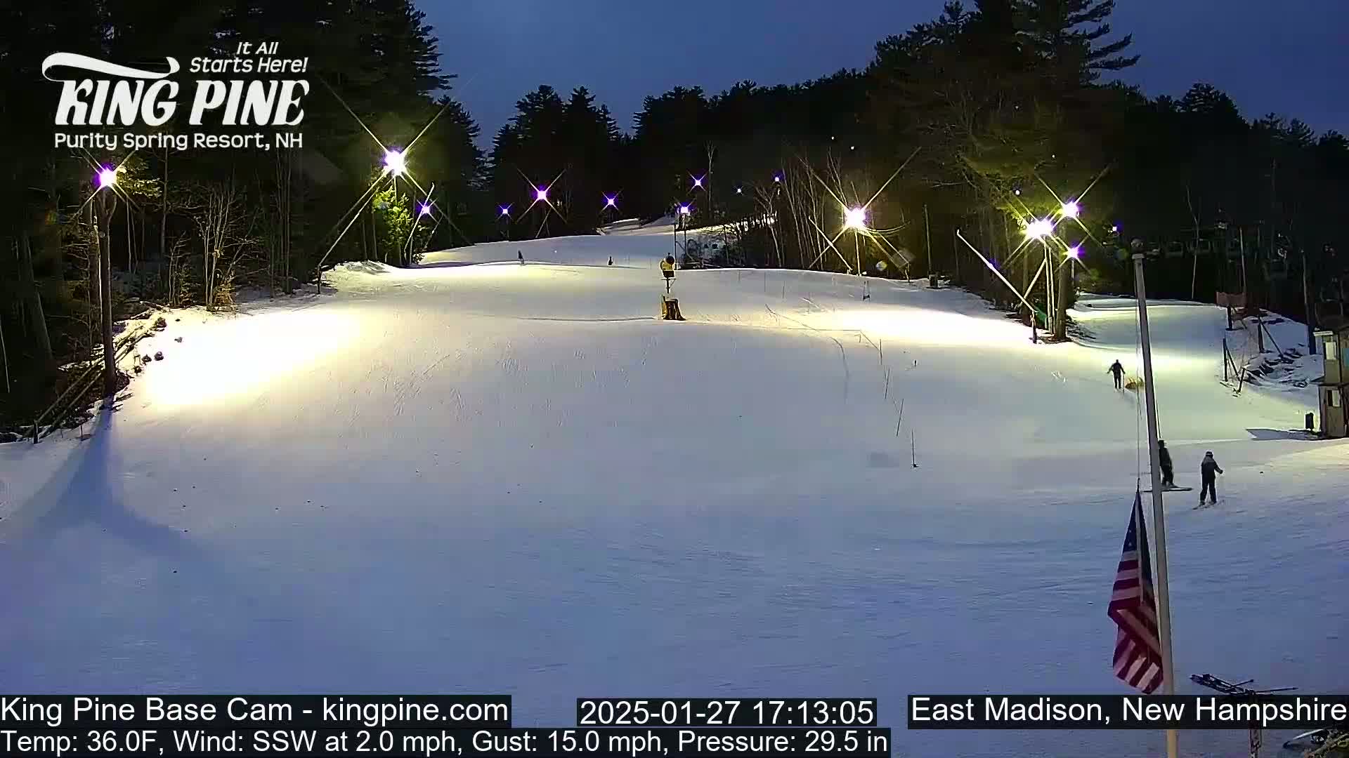 A snow-covered ski slope is illuminated at night by several lights, with a few skiers visible, under a dark sky.