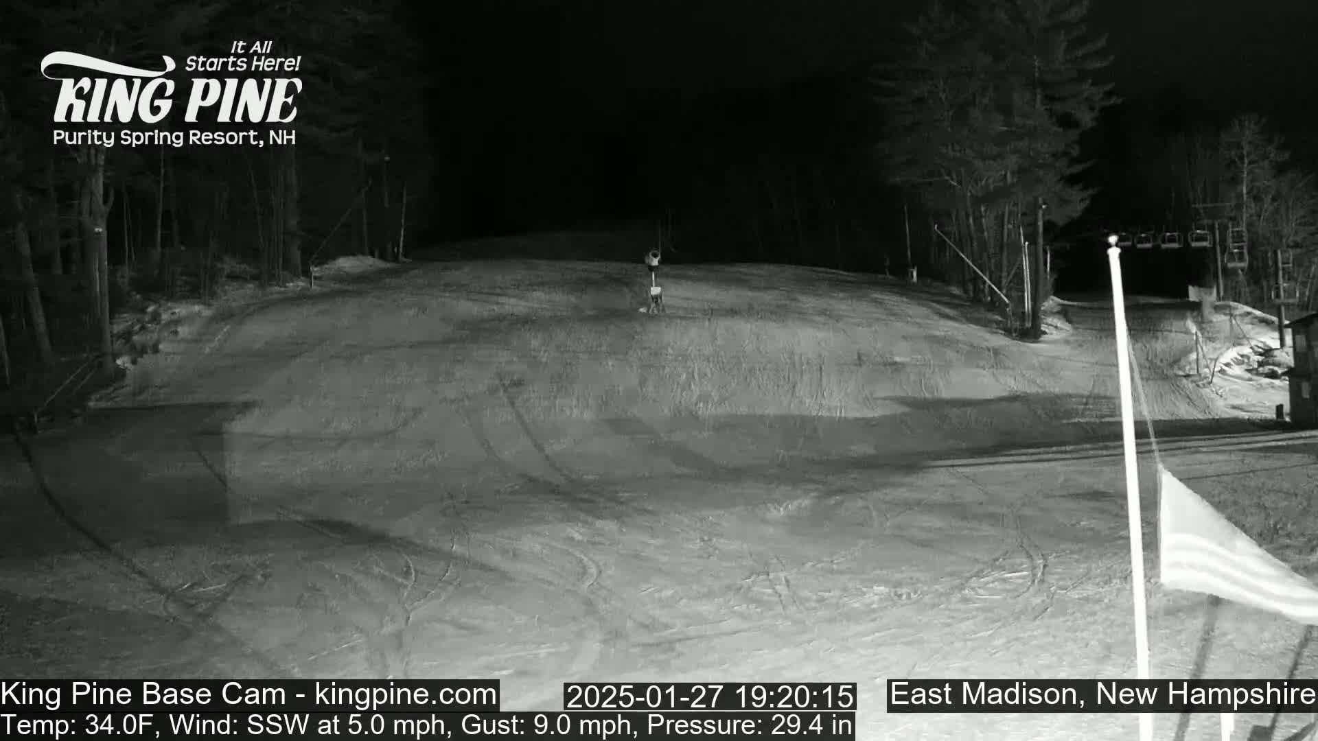 A nighttime, snow-covered ski slope is shown under a dark sky with a slight wind.