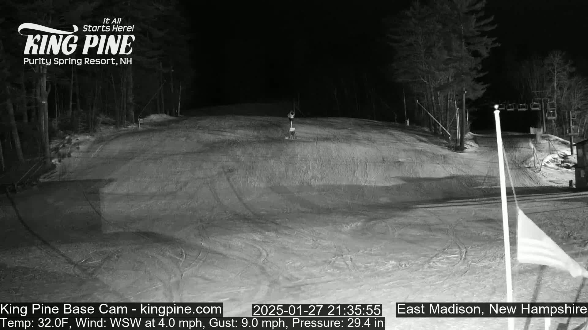 A snow-covered ski slope at night, illuminated under artificial light, with a chairlift visible in the background and a flagpole with a partially visible flag in the foreground.