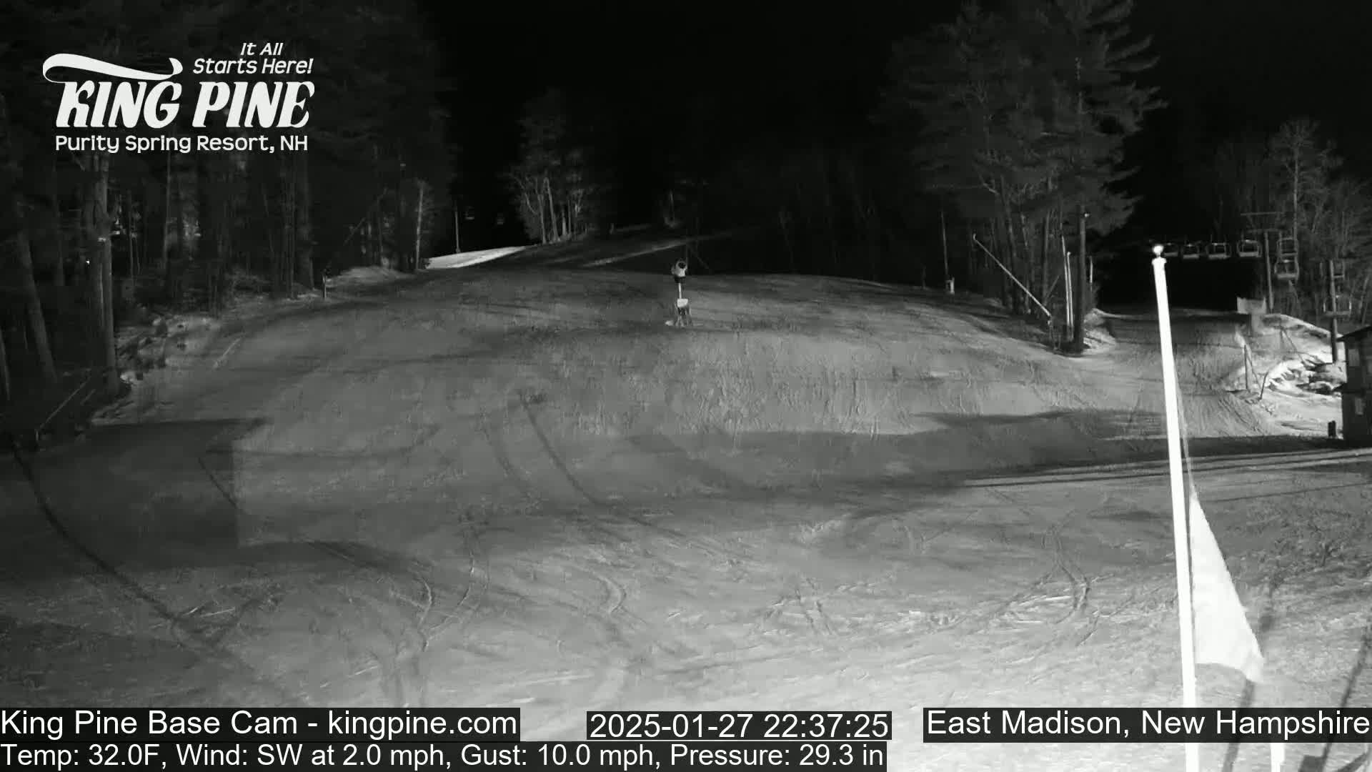 A nighttime view of a snow-covered ski slope with trees in the background, under a clear sky, with a light southwest wind.
