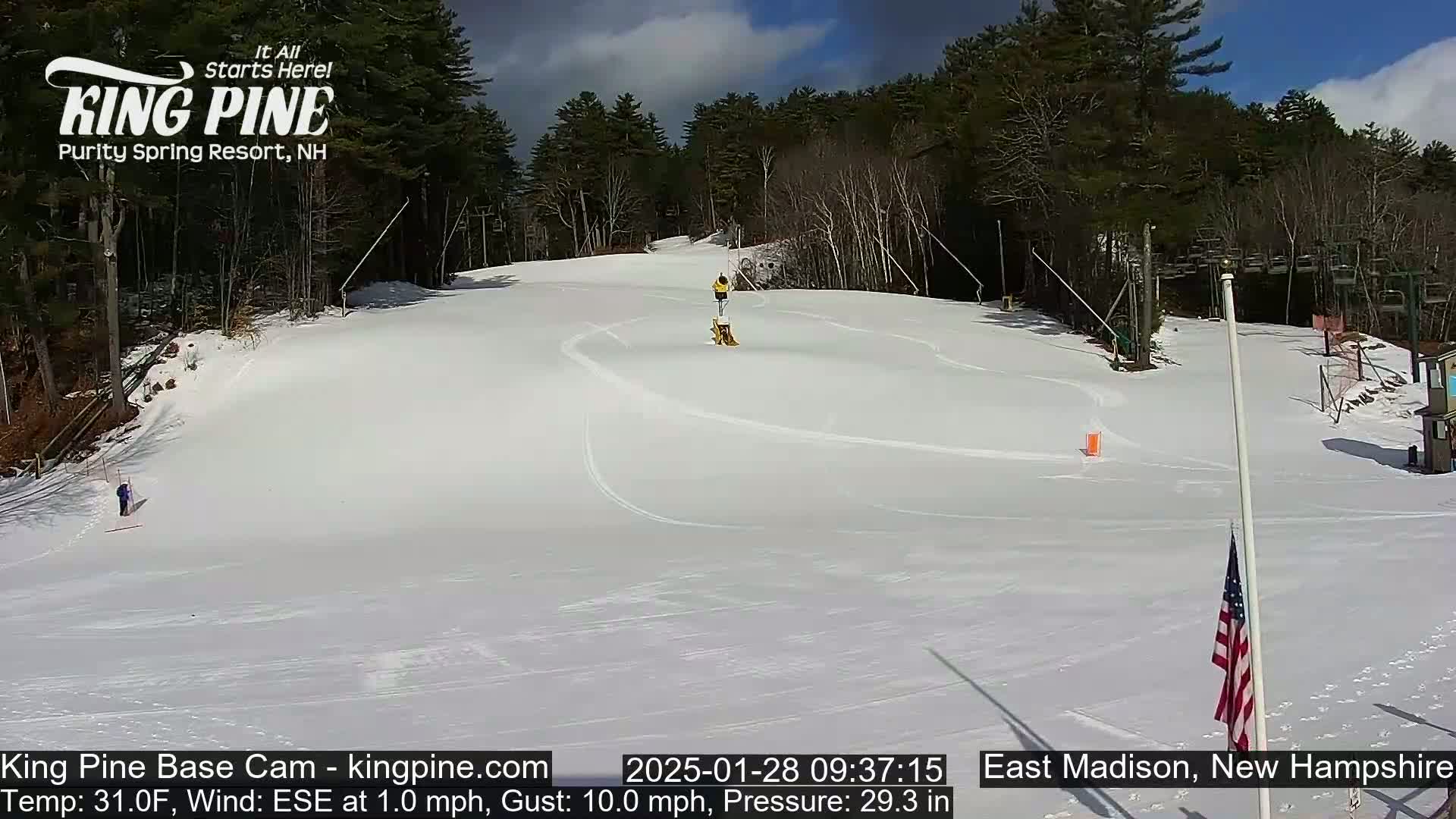 A snow-covered ski slope with a person skiing down it, under a mostly sunny sky.