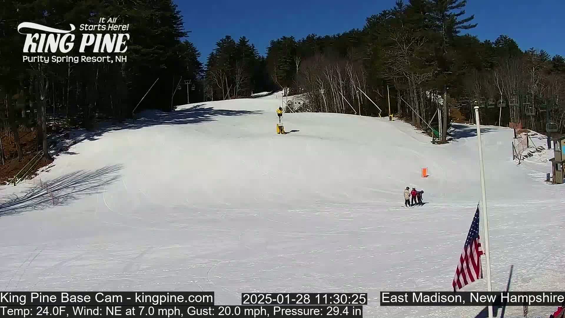 A snow-covered ski slope on a sunny day, with several people skiing or standing near the bottom, and a ski lift visible in the background.