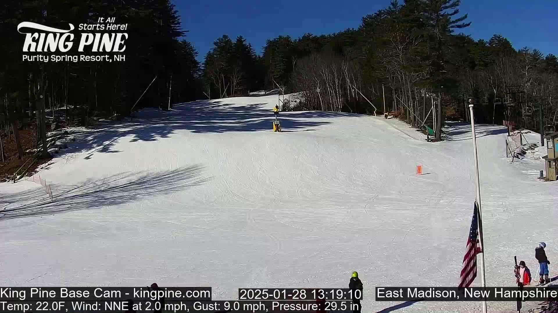 A snow-covered ski slope is visible under a clear blue sky, with several skiers and a flag visible on the lower right.