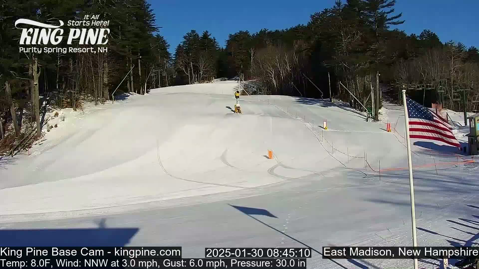 A snow-covered ski slope is visible on a sunny day, with trees lining the background and an American flag displayed on the right.