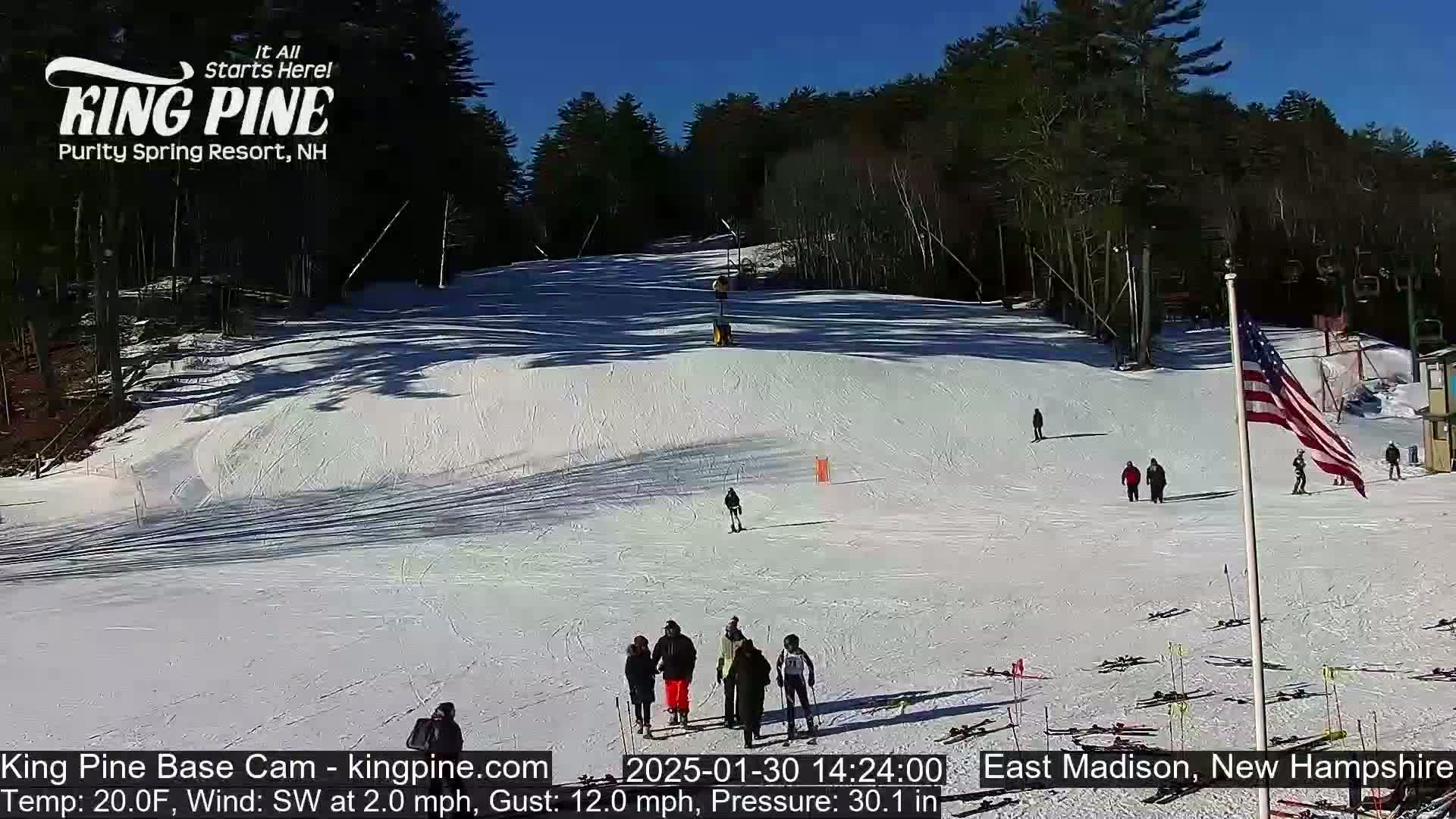 A snow-covered ski slope with several people skiing and walking, under a sunny sky.