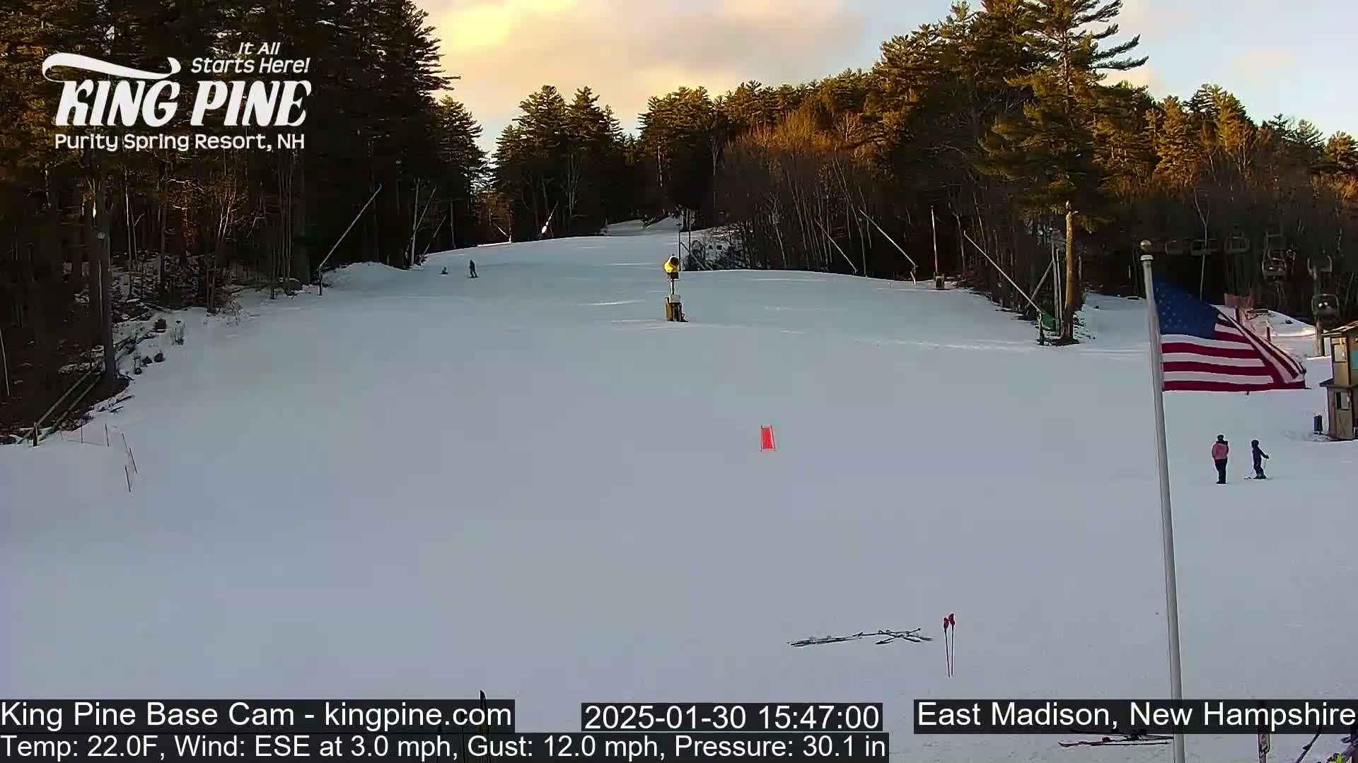 A snow-covered ski slope at sunset, with a few skiers visible and a snowmaking machine in the middle ground, is surrounded by a wooded area.