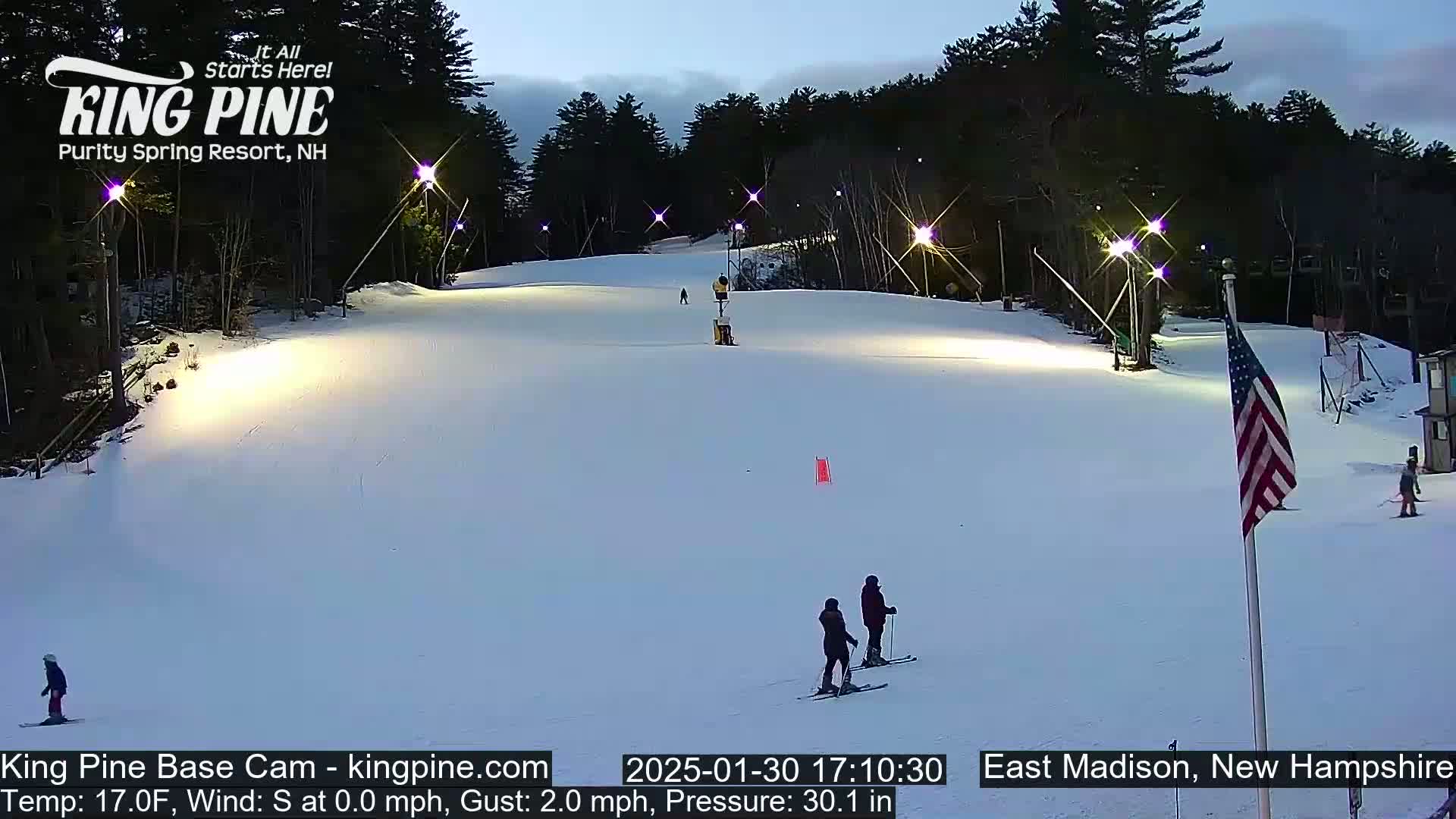 A snow-covered ski slope at dusk is lit by several bright lights, with several skiers visible on the slope.