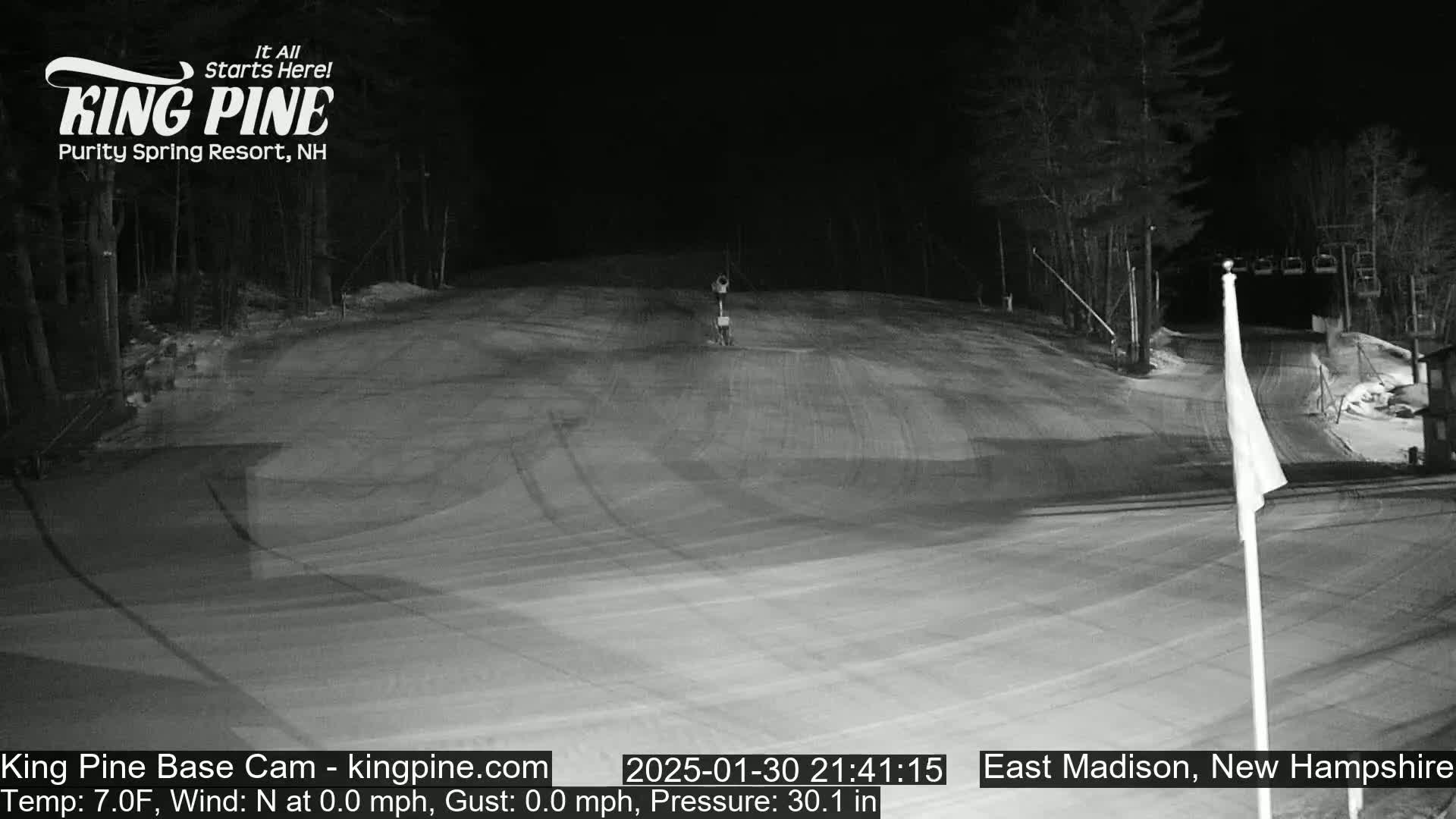 A nighttime view of a snow-covered ski slope with trees and a ski lift in the background, under calm conditions.