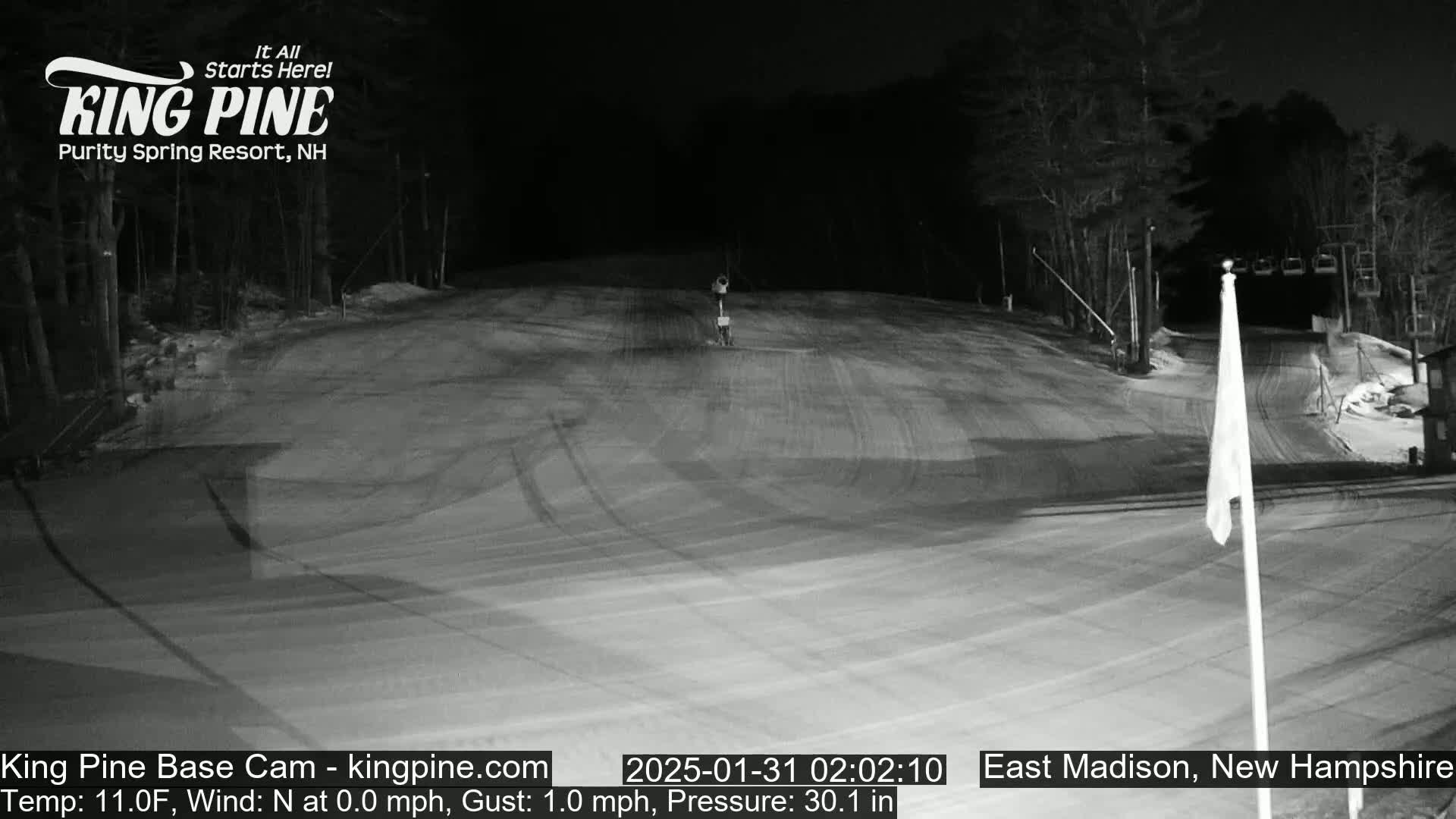 A nighttime view of a snow-covered ski slope with a chairlift and trees in the background under a dark sky.