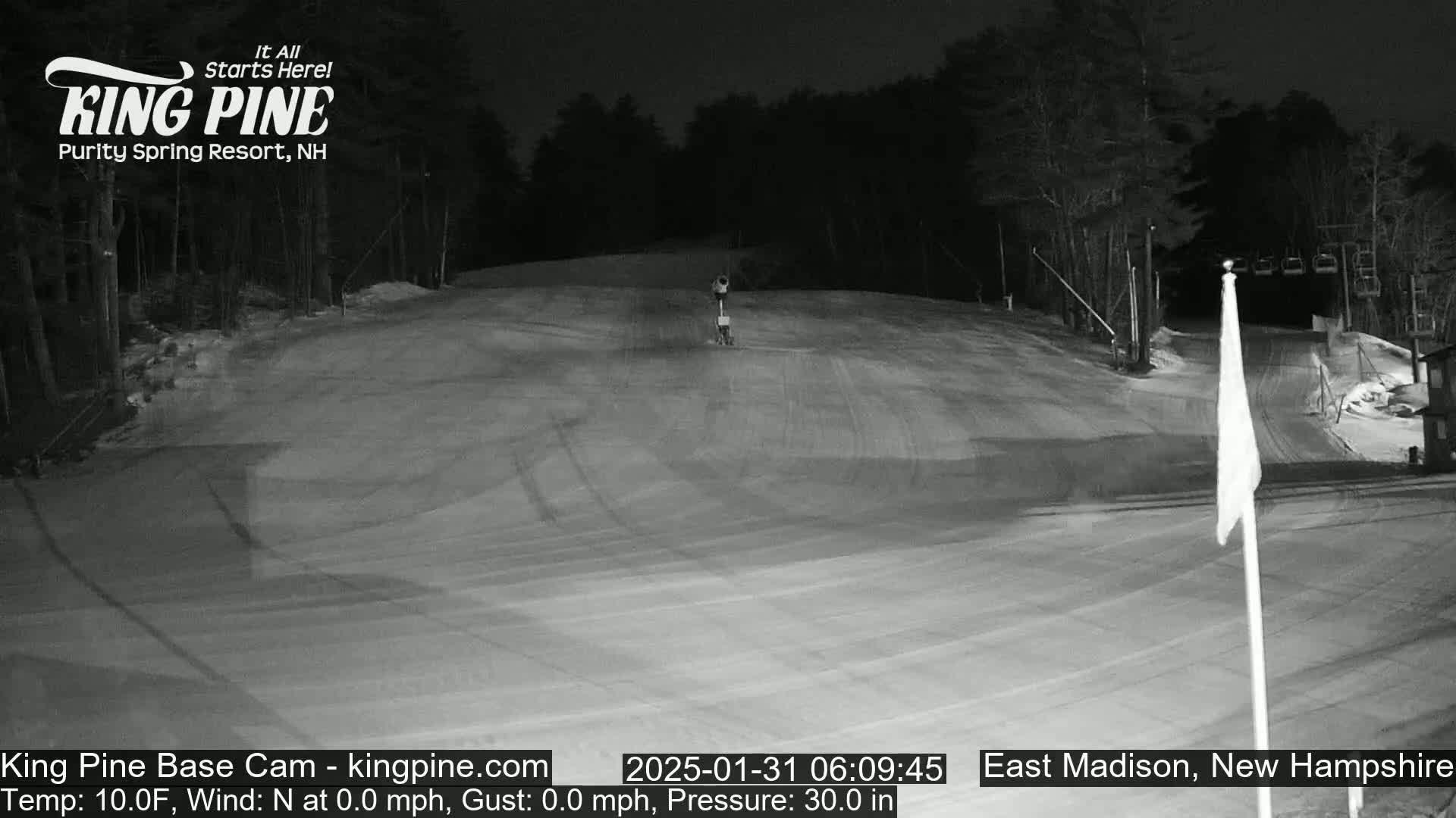 A snow-covered ski slope at night, illuminated by artificial light, with a chairlift and flag visible in the background.
