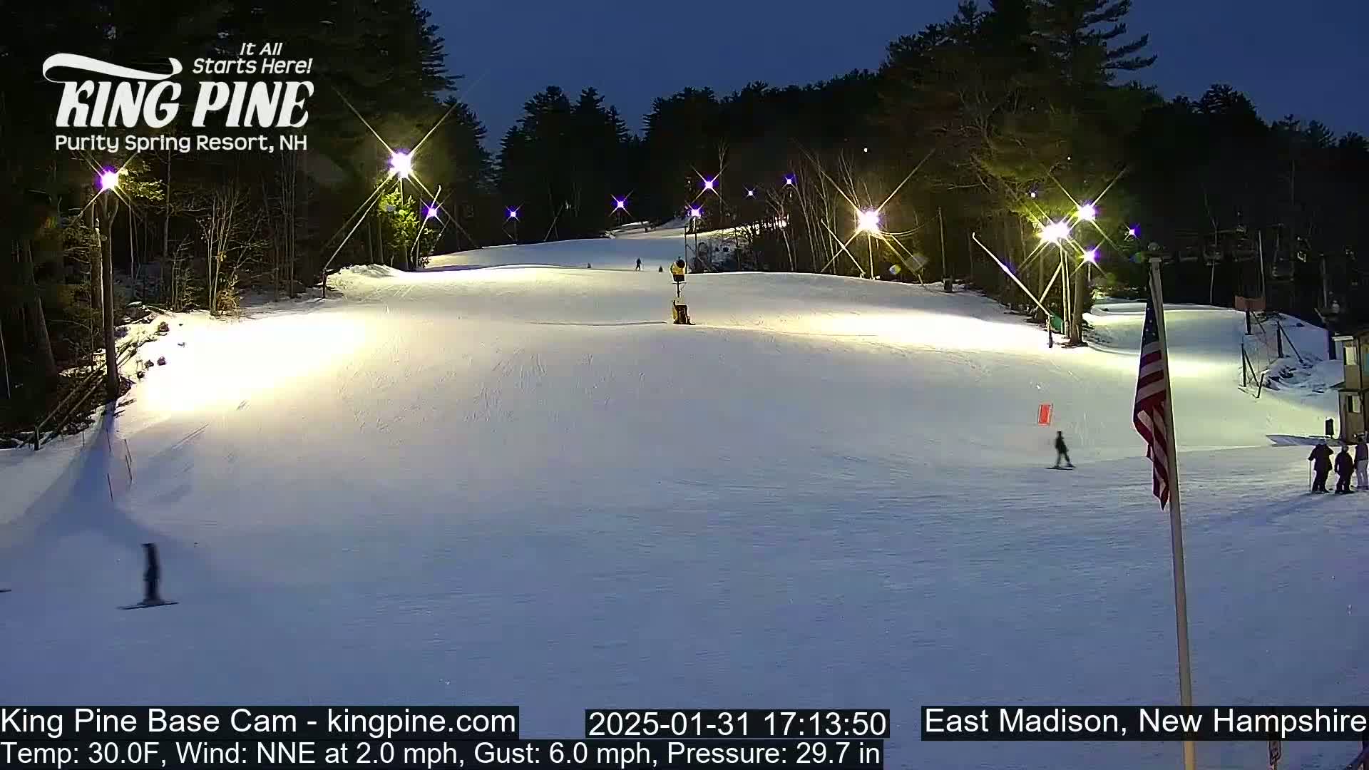 A snow-covered ski slope is illuminated at night by several lights, with a few skiers visible on the slope and a US flag standing near the bottom.