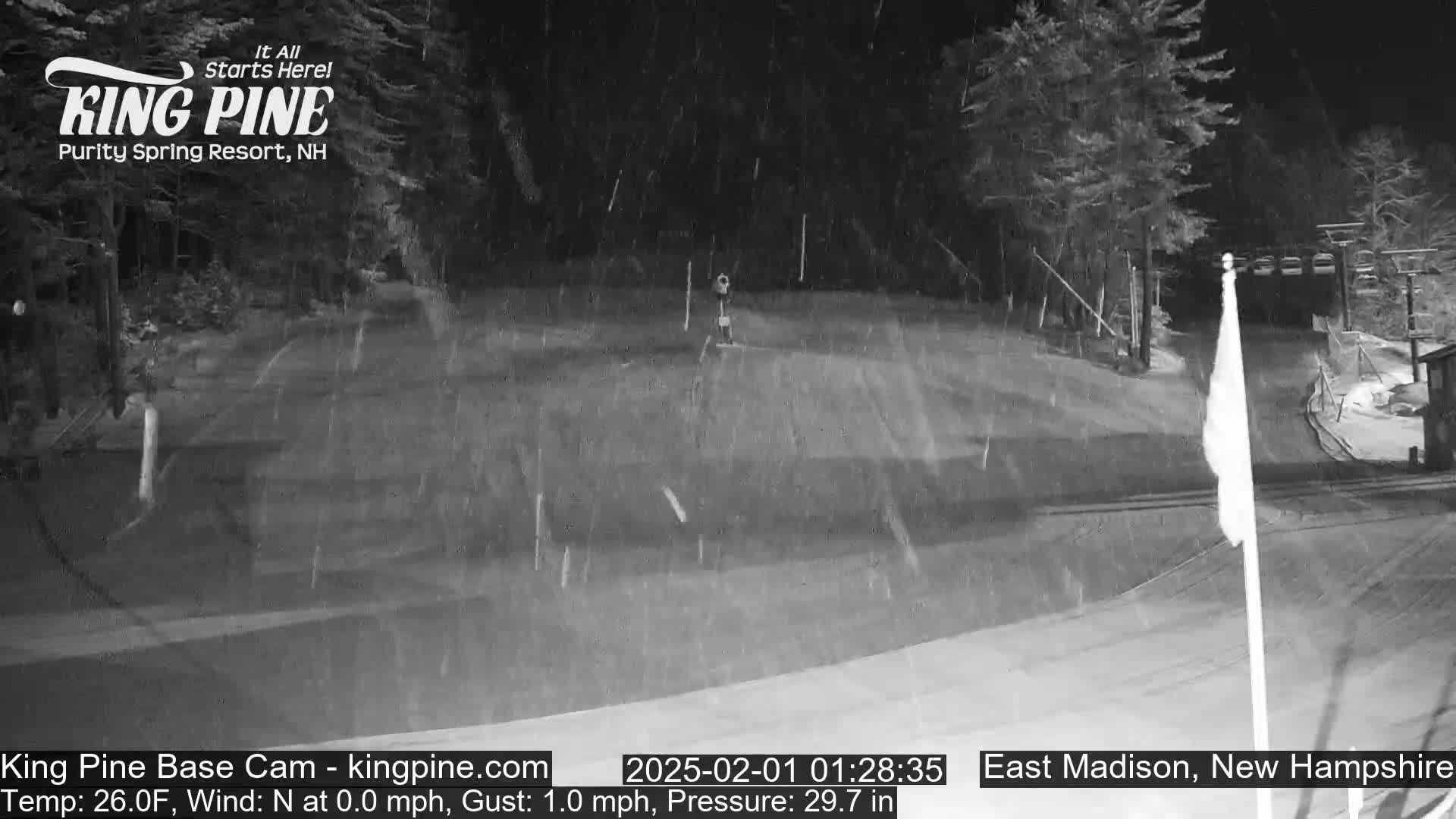 A nighttime snow scene shows a snowy, flat area with trees and a flagpole in the background under falling snow.