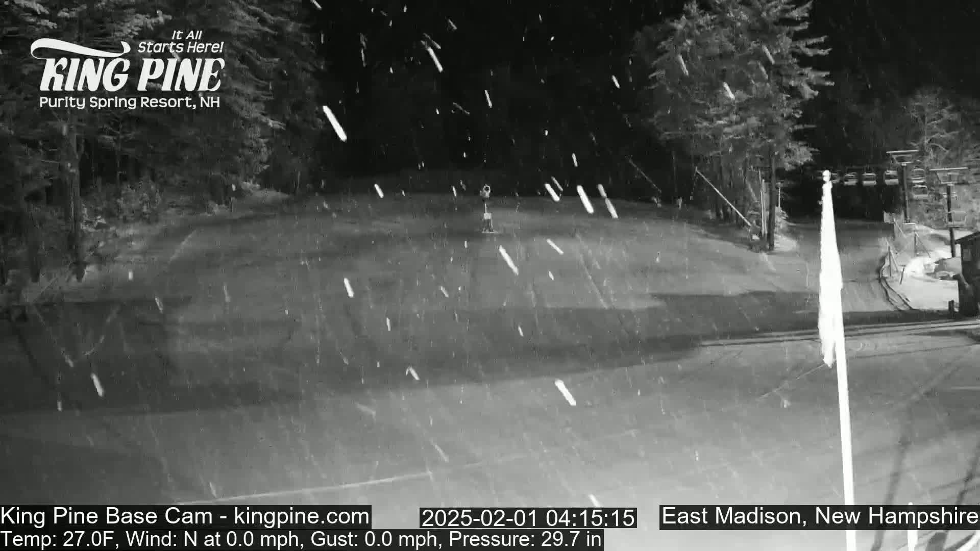 A nighttime, snowy scene shows a mostly empty paved area with a flagpole and trees in the background.