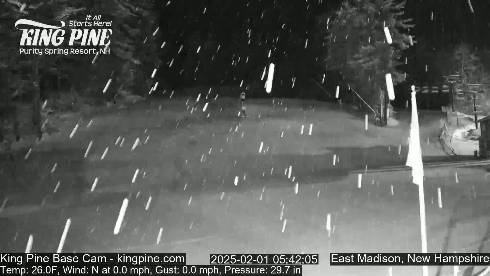 A nighttime snow scene shows a mostly empty paved area with some light snowfall.