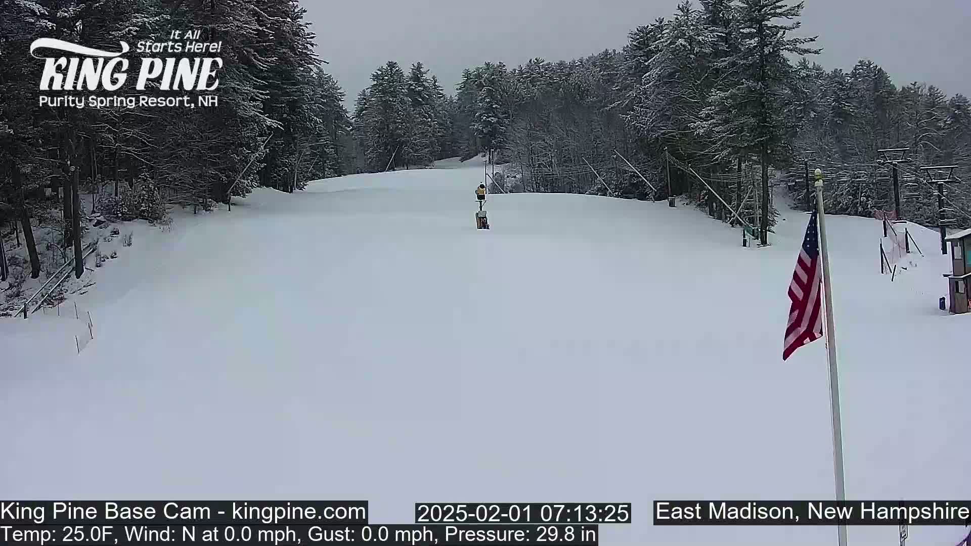 A snow-covered ski slope with a person standing near a lift, surrounded by snow-laden trees under an overcast sky.